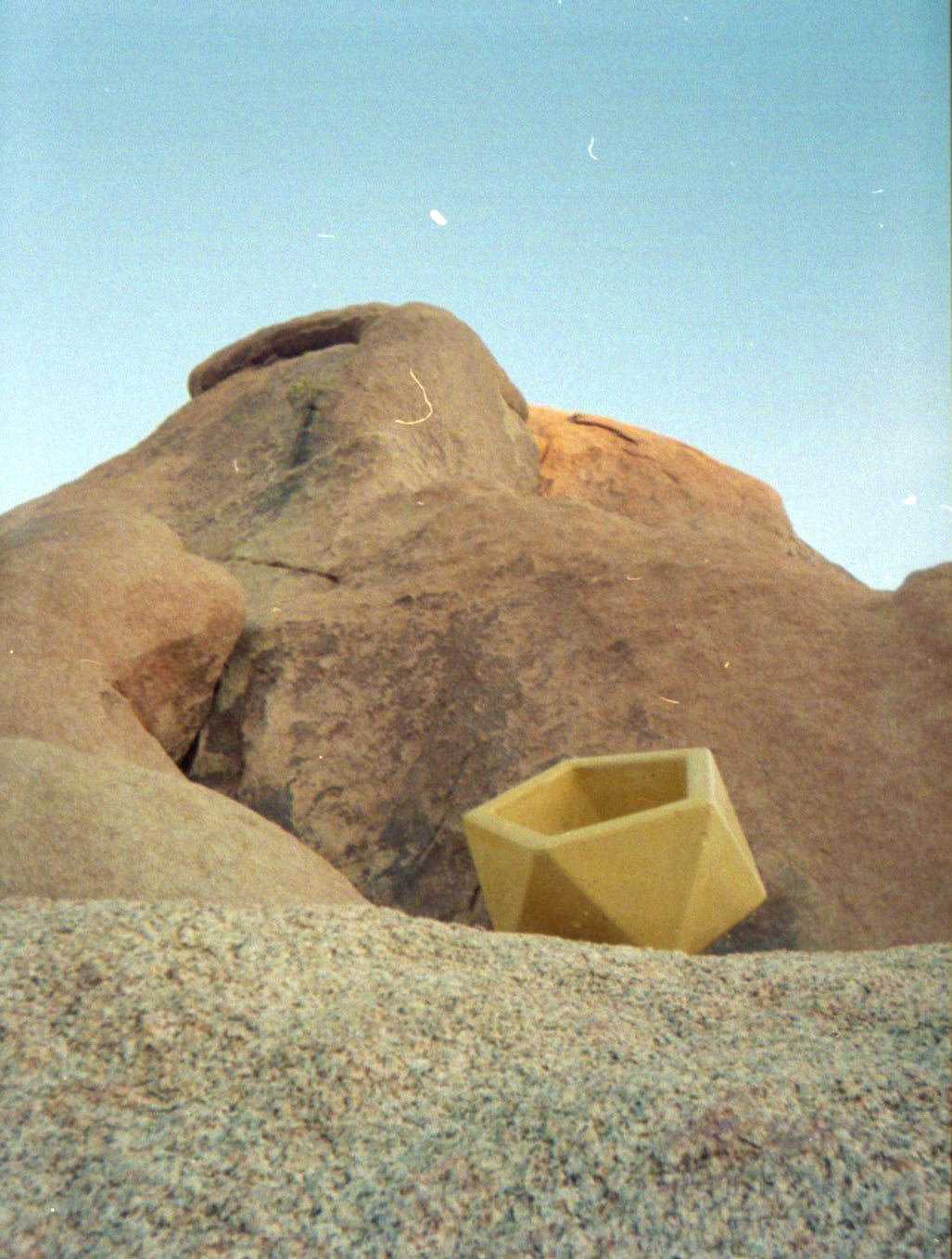 A yellow geometric concrete planter pot placed in a sandy desert landscape with large brown rock formations and a blue sky background.